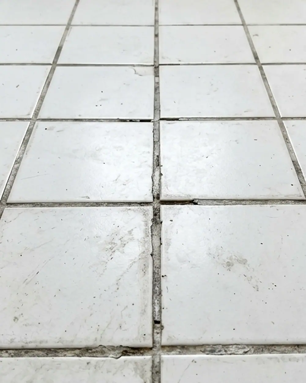 White floor tiles whose grout joints have gone grey, stained and crumbling
