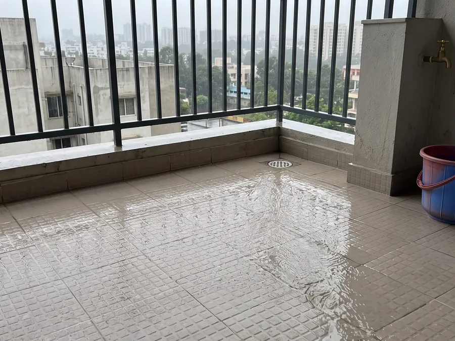A tiled apartment balcony after rain, water running across the joints toward a corner floor drain